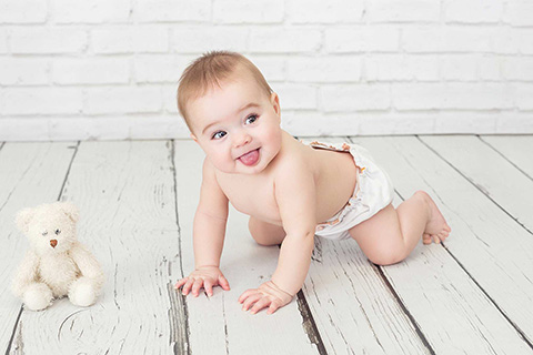 Imagen de un bebé en una sesión de fotos de un año, está en el suelo boca abajo apoyando sus manos y sus pies, lleva una braguita blanca y está con la lengua fuera mirando a cámara.