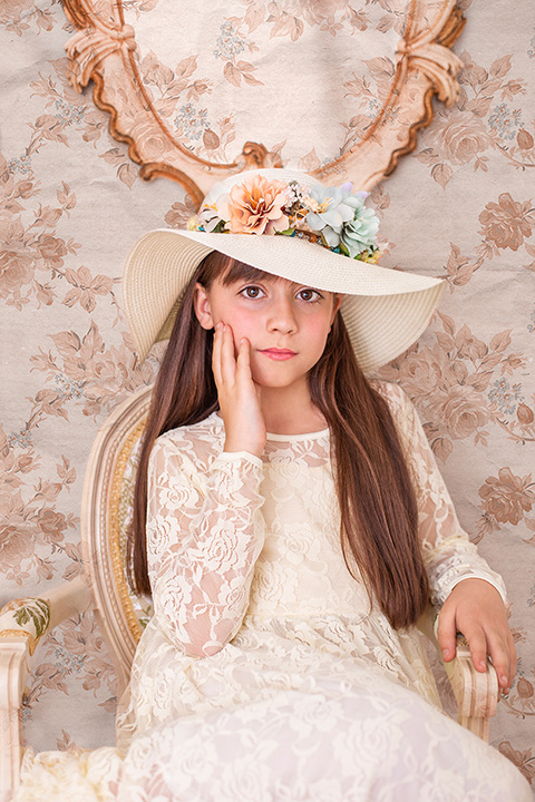 Fotógrafo de comunión en Madrid realiza una sesión de una niña morena de pelo largo, con vestido y pamela en color beige, está sentada en una silla de madera y el fondo de la imagen tiene un papel de flores y un espejo.