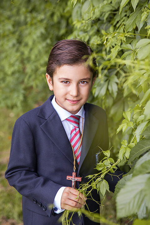Niño en su reportaje de primera comunión en exteriores, lleva una chaqueta azul, camisa blanca y corbata, de su cuello cuelga una cruz de madera y está sobre un fondo de hojas verdes.