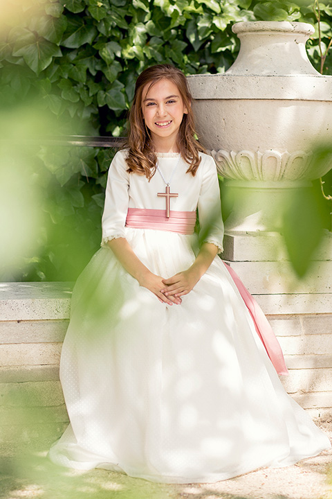 Sesión de fotos de primera comunión en exteriores, la niña se encuentra sentada en la Rosaleda del Parque del Oeste de Madrid, lleva un vestido blanco con un cinturón rosa y sobre su cuello cuelga una cruz de madera.