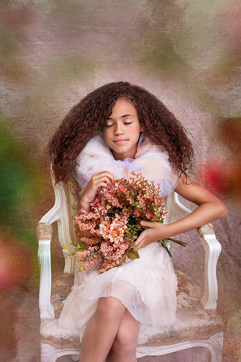 Niña pelirroja de pelo rizado sentada sobre una silla de madera blanca, lleva un vestido rosa y un cuello de tul, sujeta unas flores entre sus manos y está con los ojos cerrados en su sesión fotos de primera comunión.