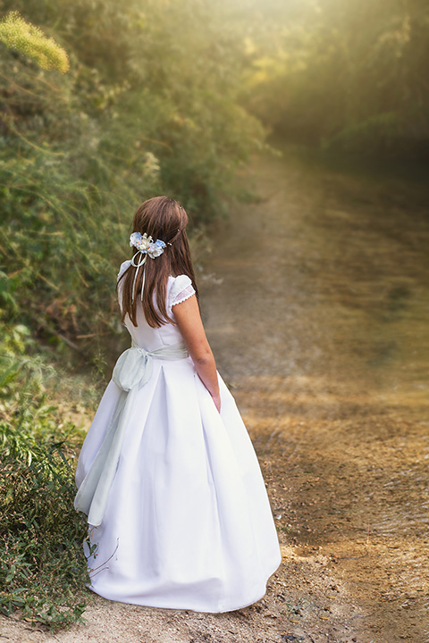 Imagen de una niña sobre un río, está de espaldas, lleva un vestido blanco con un lazo y un tocado en el pelo con flores en un reportaje de comunión en exterior.