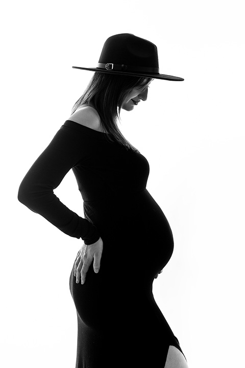 foto en blanco y negro a contraluz de mujer con vestido negro y sombrero, realizándose una sesión de embarazada en estudio de fotografía.