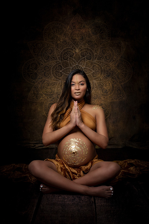 mujer sentada en el suelo en sesión de fotos para embarazada con un dibujo en la tripa en pan de oro, está en posición de yoga, con las manos en el pecho, vestida y maquillada en tonos dorados.