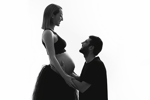 fotografía de embarazada con pareja en blanco y negro, la mujer se encuentra de pie con un top y falda de tul negra y el chico está de rodillas tocando su barriga y mirando hacia ella.