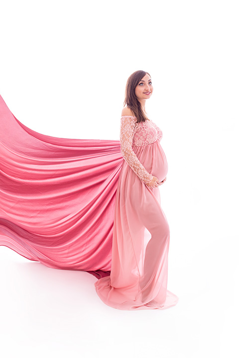 mujer en fondo blanco con un vestido gaseoso trasparente de color rosa y con una cola muy larga, muestra su silueta a una fotógrafa profesional de embarazo que no sale en la imagen.