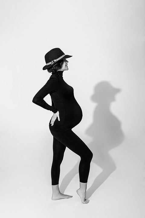 mujer embarazada vestida con un mono negro que cubre todo su cuerpo y un sombrero, está posando muy estirada y genera una sombra en el suelo con su silueta. 