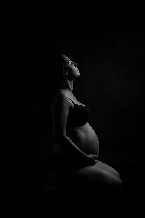 Imagen de embarazada en ropa interior en blanco y negro, es un contraluz donde se muestra el contorno de la mujer que se encuentra de rodillas mirando hacia arriba.