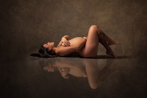 Mujer tumbada con los ojos cerrados, boca arriba con las piernas flexionadas, tapa su pecho con la mano izquierda pues se está realizando una fotografía de desnudo en estudio de embarazo.