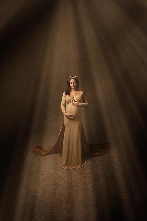 Fotógrafo de embarazadas en Madrid captura la imagen de una mujer con vestido dorado con cola y diadema del mismo color, ella mira hacía la cámara y toca con sus dos manos su tripa.