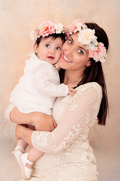 Madre e hija en una sesión de fotos familiar en estudio, están abrazadas y llevan un vestido beige y coronas de flores la cabeza en tonos rosas y verde agua.