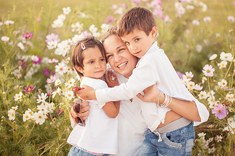 Madre e hijos están sentados sobre un campo de flores blancas y moradas, los niños abrazan a su madre y sonríen, llevan ropa blanca y vaquero, es una fotografía de familia en exteriores.