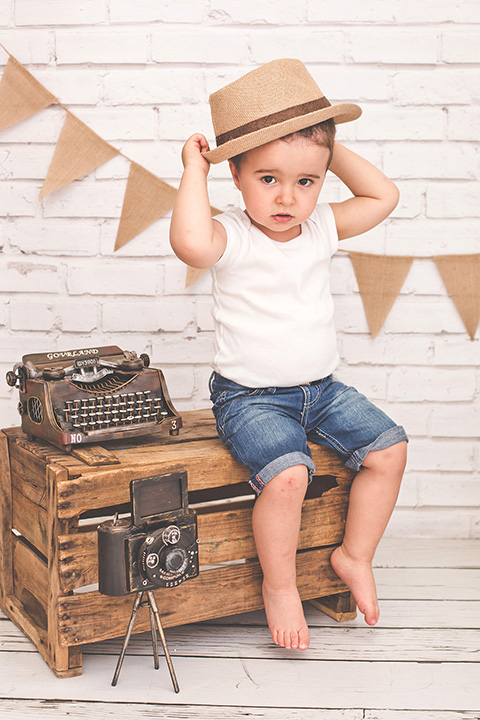 Niño sentado sobre una caja de madera con camiseta blanca, vaquero y sombrero. Tiene una máquina de escribir y una cámara de fotos antigua, la imagen tomada en una sesión infantil en estudio.