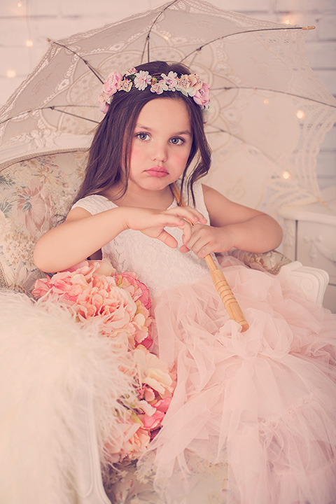 Niña en fondo blanco con vestido de tul rosa y una corona de flores, está sentada sobre una silla y está sujetando un paraguas en un book de fotos infantil en Madrid.