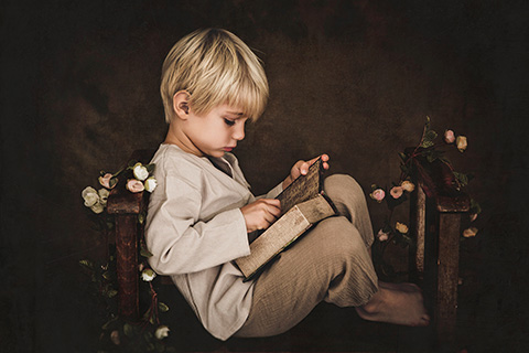 Books de fotos para modelos infantiles de un niño rubio sentado sobre un banco con flores y leyendo un libro antiguo, lleva una camisa beige y un pantalón marrón.