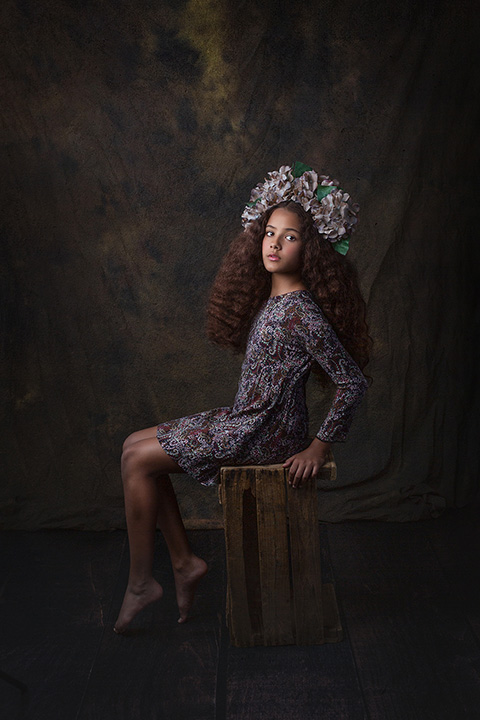 Niña de pelo rizado con flores en la cabeza sentada sobre un cajón de madera en unas fotos para firma de moda infantil.
