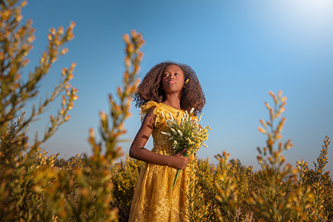 Sesiones de fotos infantiles en exteriores, niña de pelo rizado con vestido amarillo en campo de flores, sujeta varias entre sus manos.