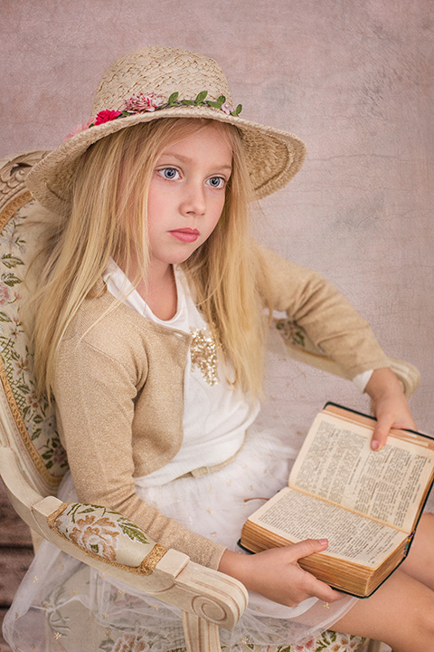 Niña rubia de pelo largo, sentada sobre una silla de madera beige con flores, lleva un sombrero y una chaqueta, sujeta un libro abierto entre sus manos realizada por un fotógrafo de niños en Madrid.