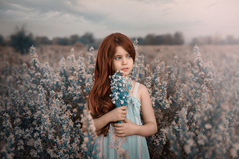 Catálogo de moda infantil en exterior de niña pelirroja de pelo largo en campo de flores blancas y verde agua, con vestido a juego, también sujeta un ramo entre sus manos.