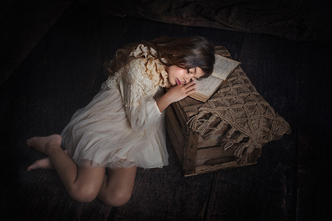 Foto de una niña con los ojos cerrados tumbada sobre un cajón de madera, lleva un vestido corto beige y tiene la cara y las manos apoyadas sobre un libro antiguo en una sesión fine art en estudio.
