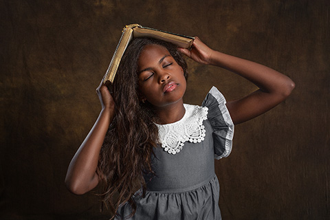 Imagen de una niña con pelo largo y un vestido gris con cuello blanco, tiene un libro en su cabeza que sujeta con sus manos, es una fotografía infantil en Madrid.