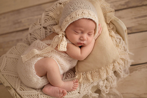 Sesión newborn de un bebé tumbado en una cama pequeña blanca, viste un body y gorro beige de encaje.