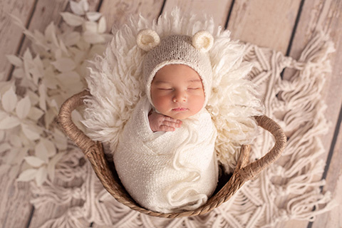 Bebé envuelto en una tela blanca y lleva en su cabeza un gorro lana con forma de oso, está metido en una cesta de mimbre sobre una alfombra de crochet.