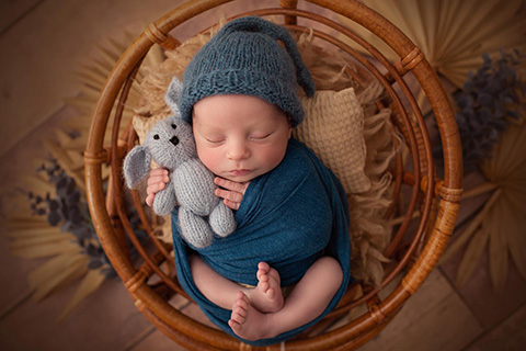 Foto newborn de un niño dormido, tambado boca arriba en un soporte de ratán, envuelto en una tela azul marino con gorro de lana del mismo color, además está sujetando un conejo de peluche.