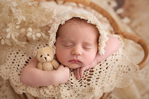 Bebé metido en una cesta de ratán, está dormido y sujeta un oso con su mano derecha, lleva un gorro beige con flecos y esta sobre una manta de crochet del mismo color.
