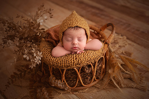 Bebé recién nacido metido en una cesta de bambú, esta boca abajo apoyada su cabeza sobre sus manos, lleva un gorro tono tostado y está sobre una manta del mismo color.
