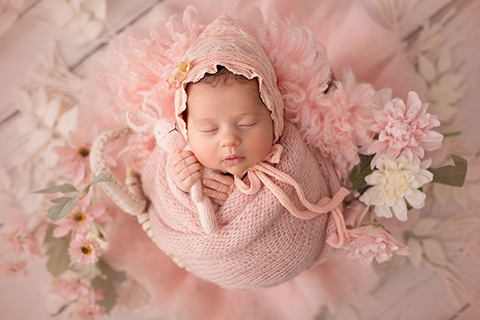 Bebé niña recién nacida envuelta con una tela de lana rosa, lleva un gorro a juego y sujeta un peluche con sus manos.