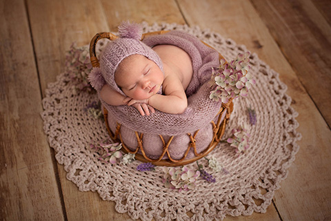 Bebé con gorro morado con pompones, en una cesta de bambú con flores a su alrededor y sobre una manta redonda de ganchillo.