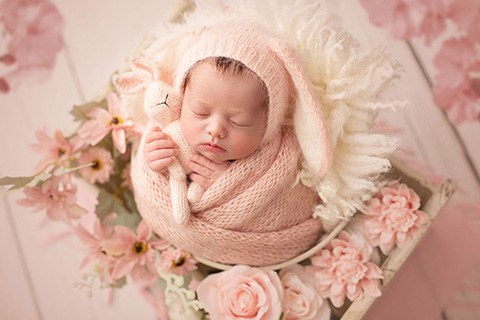 Preciosa bebé con gorro de conejo en color rosa, duerme en un cajón de madera lleno de flores.