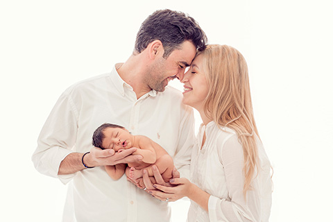 Padre y madre sujetan con sus manos a su bebé en sus primeros días de vida, ellos tienen sus frentes apoyadas y sonríen, llevan camisas en color blanco, imagen realizada por por una fotógrafa de recién nacido en Madrid.