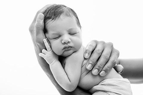 Imagen en blanco y negro de primer plano de un bebé dormido en sus primeros días de vida, está apoyado sobre la mano de su padre y la otra mano le sujeta la espalda, imagen realizada en sesiones fotográficas para recién nacido en Madrid.