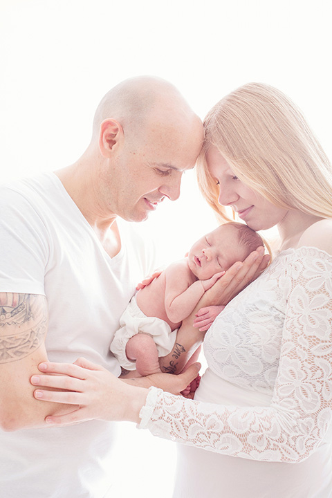 Padre y madre sujetan entre sus brazos a su bebé dormido de tan solo uno días de vida, llevan camisetas en color blanco y están sobre un fondo de estudio fotográfico especializado en recién nacido.