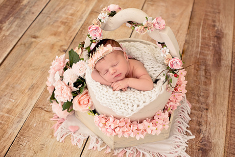 Bebé en un cubo de madera beige, está dormida y apoya su cara sobre sus manos, lleva una cinta en la cabeza y está rodeada de flores de color rosa, imagen realizada por un fotógrafo especializado en recién nacido.